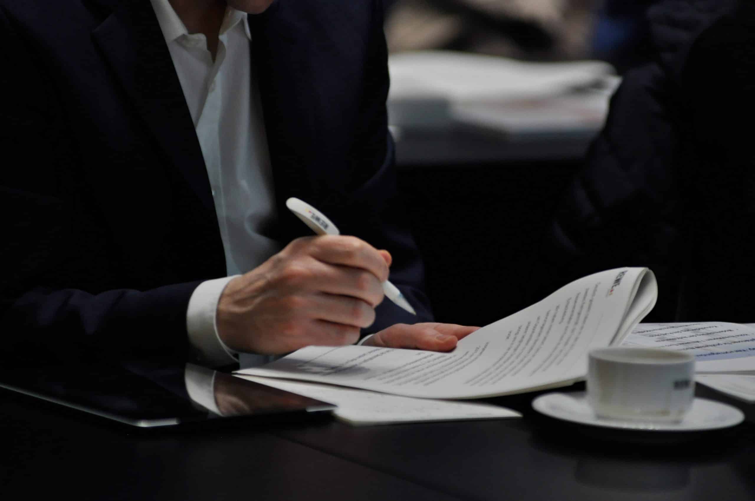 Ein Mann im Anzug hält einen REWE Group Kugelschreiber und liest dabei ein Dokument
