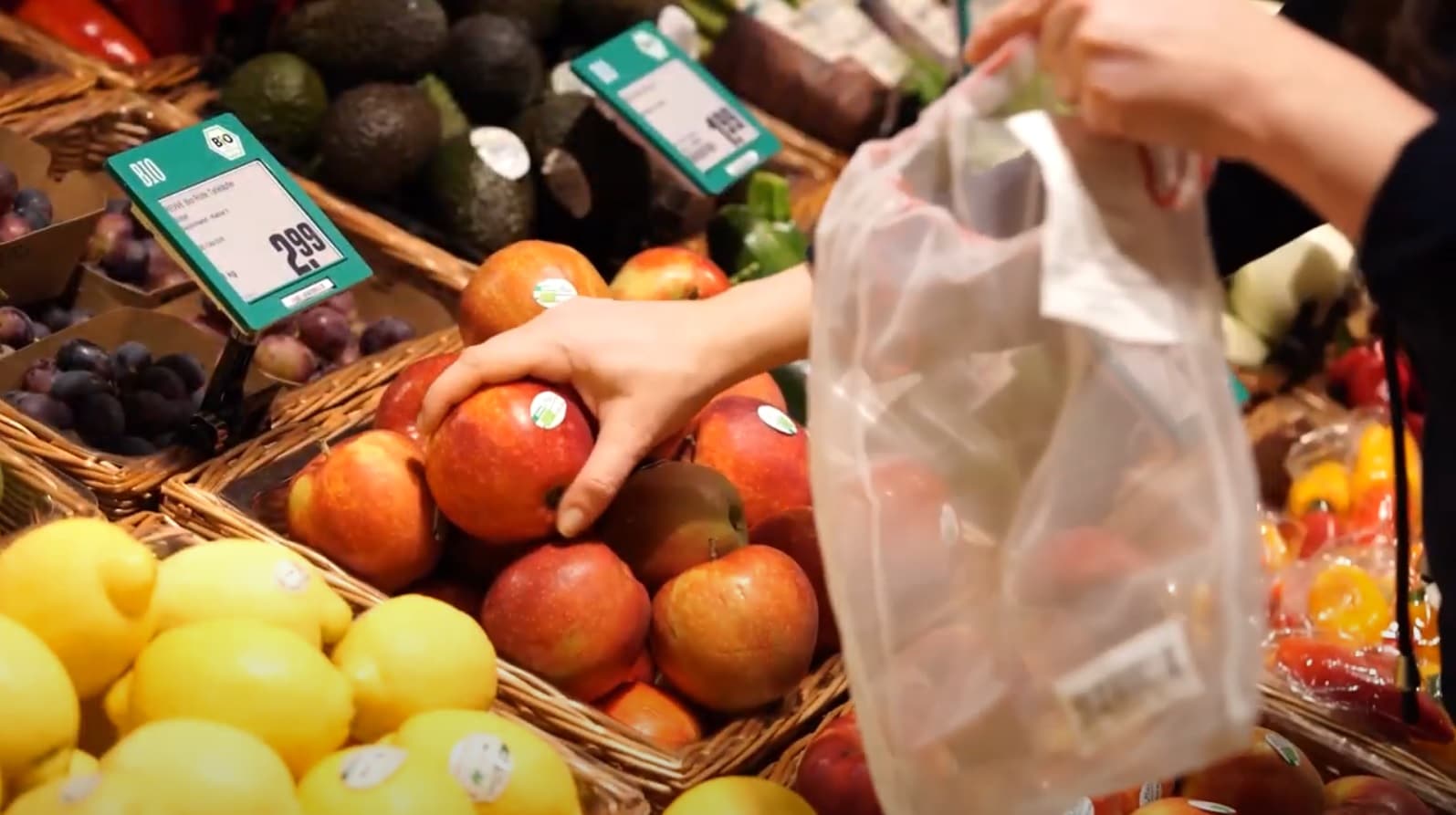 Eine Frau, die einen Apfel aus einer Korb der Obsttheke einer REWE Filiale nimmt, um dieses unverpackte Obst in eine Plastiktüte zu verpacken