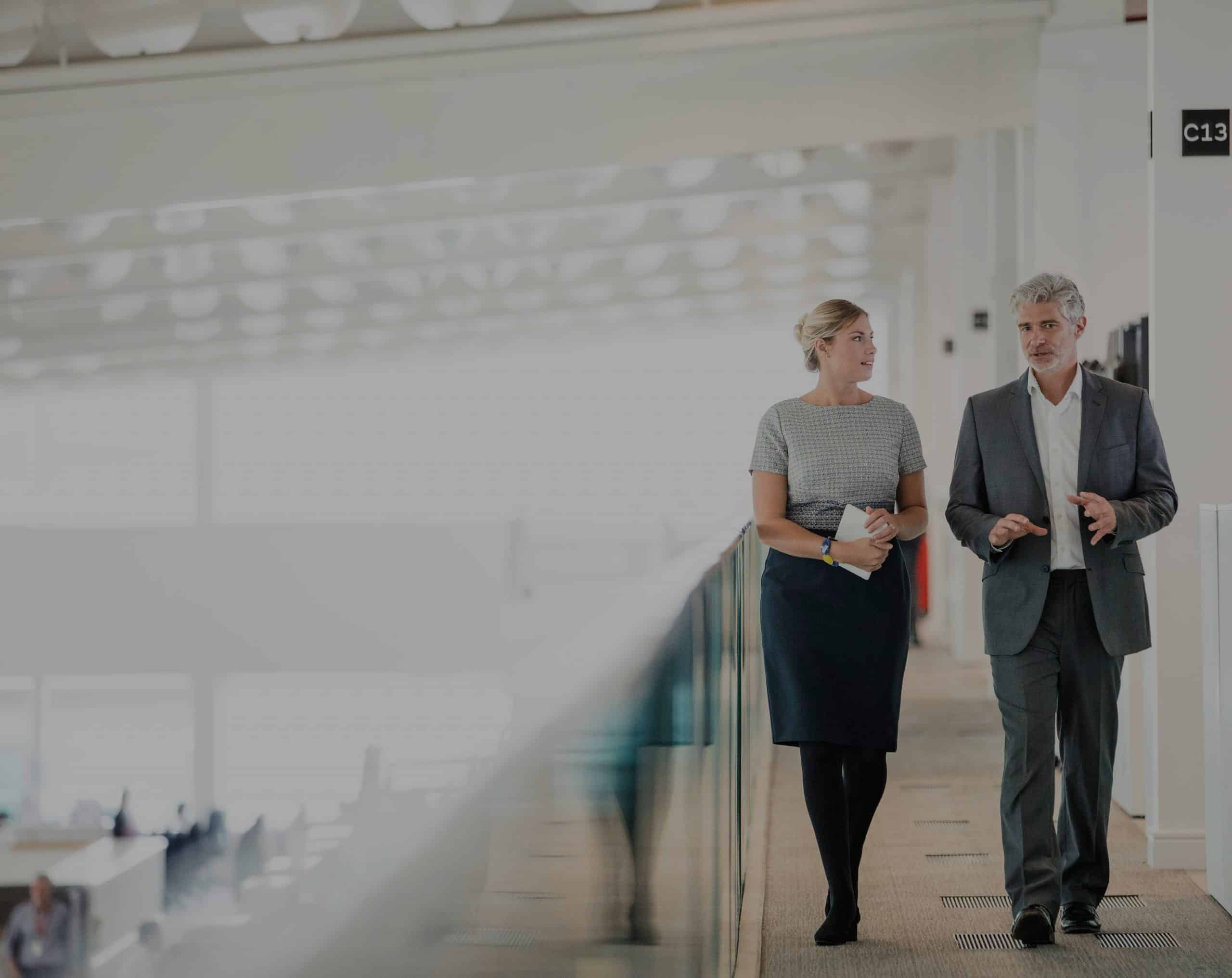 Männliche und weibliche Büroangestellte unterhalten sich, während sie über einen erhöhten Gehweg in einem Büro-Auditorium schlendern. Ein Symbolbild für den Ausichtsrat der REWE Group