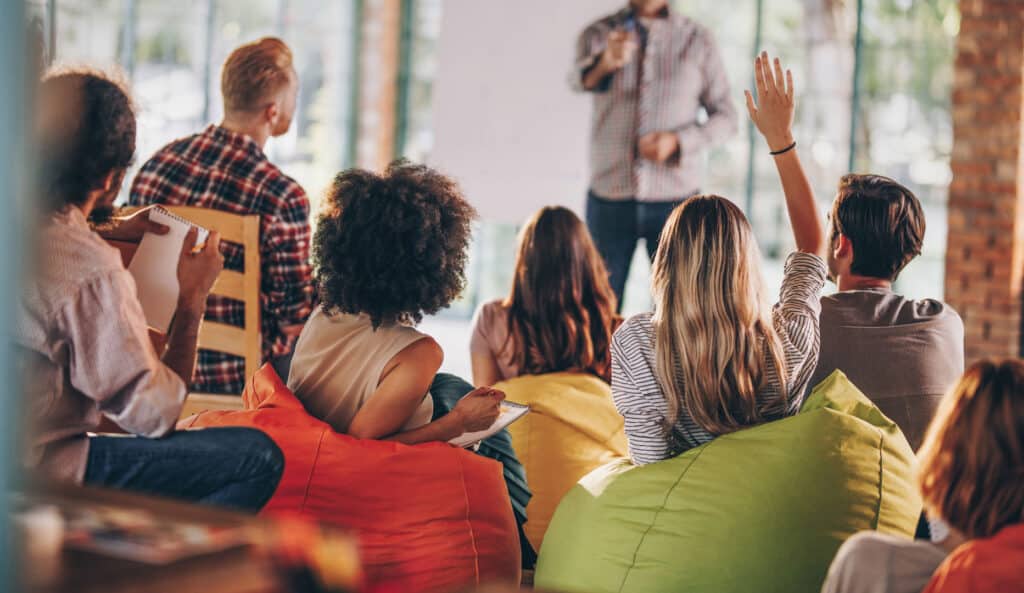 Seminarteilnehmende sitzen auf bunten Sitzsäcken, eine Teilnehmerin meldet sich mit einer Frage