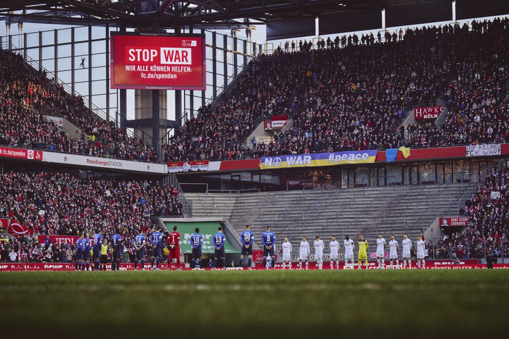Fußballmannschaft vor gefüllten Zuschauerrängen im 1. FC Köln Stadion, die vor einer Leinwand mit dem Schriftzug Stop War stehen.
