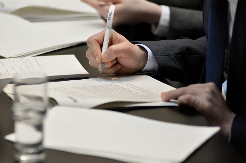 Eine Person im Anzug hält einen REWE Group Kugelschreiber und liest dabei ein Dokument