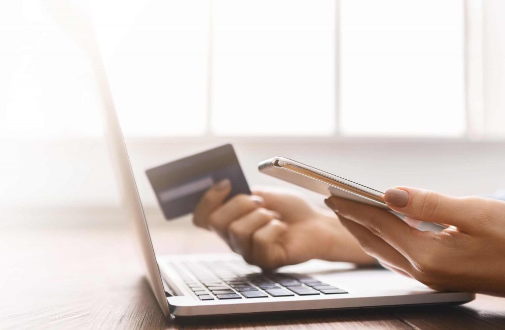 Eine Person hält eine Kreditkarte und ein Handy und sitz vor einem Laptop.