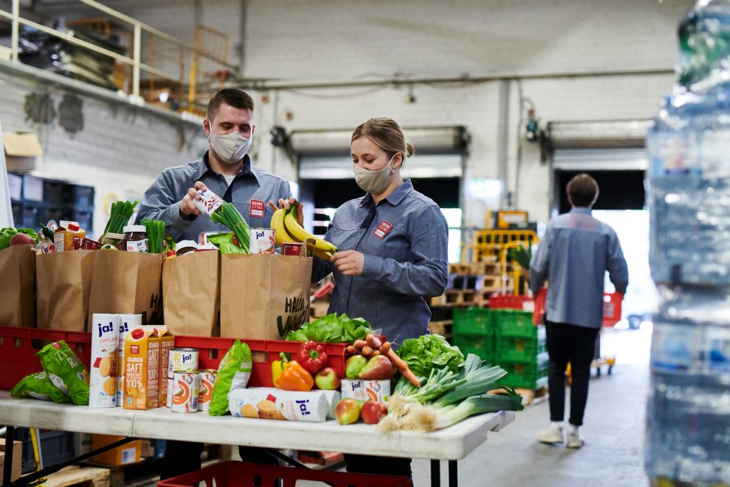Zwei REWE Mitarbeitende packen Lebensmittel in REWE Papier-Tüten